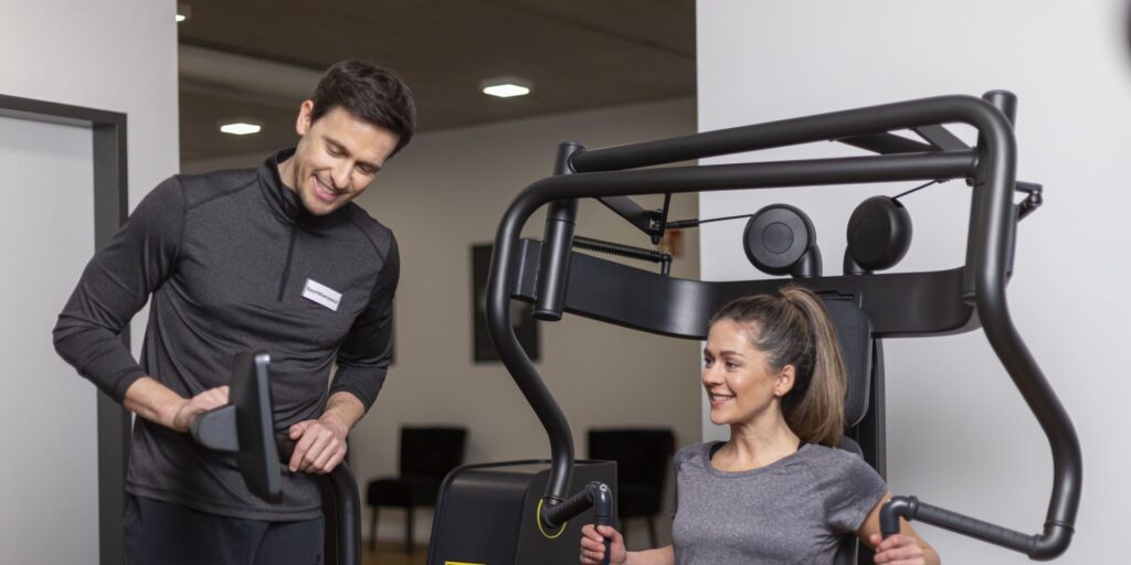 Sporttherapeut begleitet eine Frau beim Gerätetraining und überwacht die Einstellungen